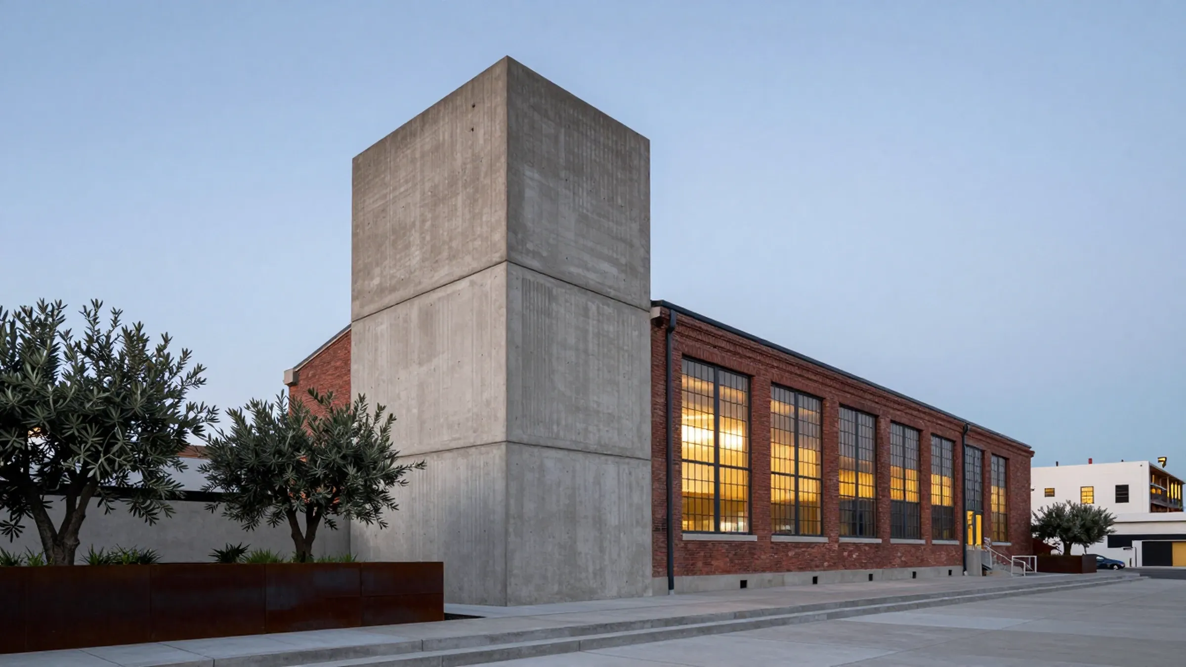 Board-formed concrete tower joined to an old brick warehouse at dusk, olive trees in front